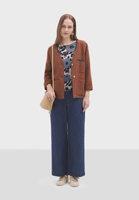 Giacca marrone con texture, dotata di due tasche frontali, indossata sopra un top floreale. Un paio di pantaloni in denim blu a gamba larga e scarpe da ginnastica beige completano l'outfit.