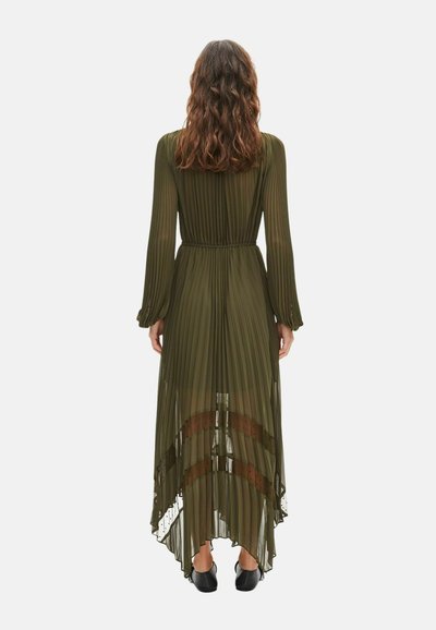 Woman with wavy brown hair wearing a long-sleeve, olive green pleated dress with sheer fabric and asymmetrical hem, viewed from the back.