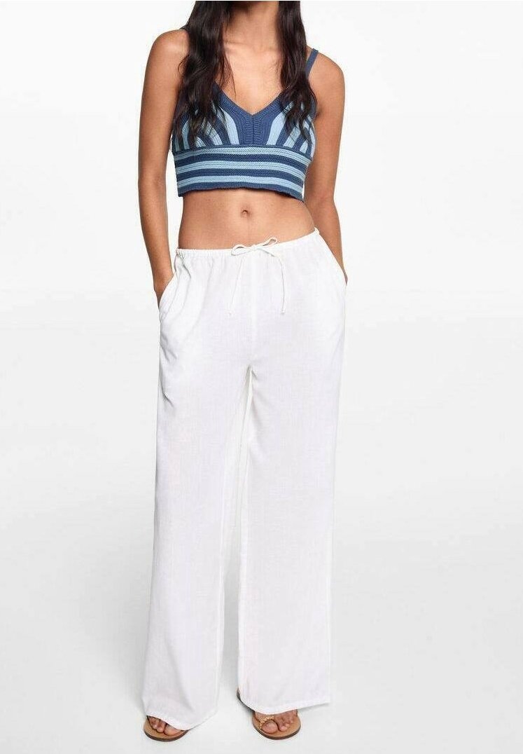 Femme portant un crop top sans manches à rayures bleues et un pantalon blanc à jambes larges avec une taille à cordon, debout les mains dans les poches.