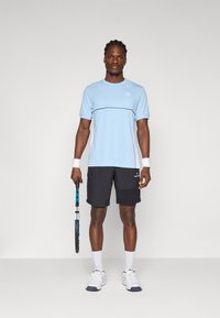 Camisa deportiva azul claro con ribetes y logo negros, combinada con pantalones cortos negros. Medias blancas y zapatillas de tenis. Sosteniendo una raqueta de tenis.