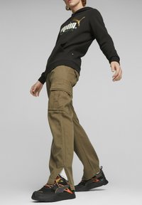 Sudadera negra con capucha y logo de Puma en verde y dorado, combinada con pantalones cargo oliva que tienen bolsillos y un dobladillo dividido, y zapatillas negras.