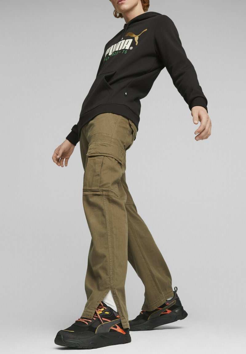 Sudadera negra con capucha y logo de Puma en verde y dorado, combinada con pantalones cargo oliva que tienen bolsillos y un dobladillo dividido, y zapatillas negras.