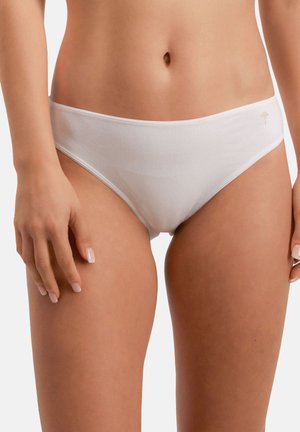 Nainen, joka käyttää yksinkertaista valkoista bikiinityylistä alusvaatetta, jossa on pieni beige logo oikeassa lonkassa, seisoo neutraalia taustaa vasten.