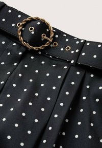 Tissu noir à pois blancs avec un design plissé et une ceinture noire avec une boucle circulaire dorée torsadée.