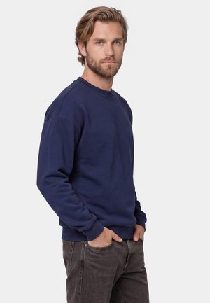 Bearded man with light brown hair wears navy blue sweatshirt and dark grey jeans with hands in pockets, facing forward on plain background.