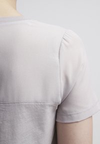 Světle fialový top s krátkým rukávem a hladkou texturou. Obsahuje na rameni nařasené detaily a barevně blokovaný design na zadní straně.