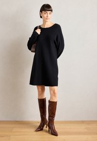 Robe noire en maille surdimensionnée à manches longues, associée à des bottes à hauteur de genou en crocodile verni marron et à un sac marron.