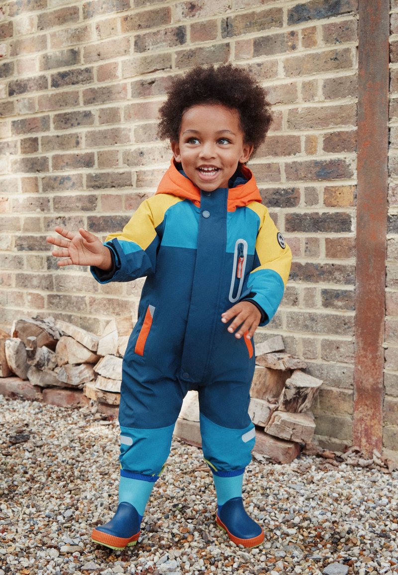 Tuta imbottita per bambini dai colori vivaci, con base navy, cappuccio arancione, sezioni gialle e turchesi, chiusura frontale con zip e dettagli riflettenti.