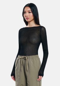 Maglia nera a maniche lunghe con un motivo trasparente e ondulato. Abbinata a pantaloni chiari con coulisse in oliva, che mostrano una vestibilità rilassata.