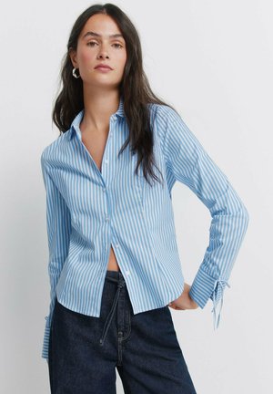 Młoda kobieta z długimi ciemnymi włosami nosi jasnoniebieską i białą koszulę w paski zapinaną na guziki oraz ciemne dżinsy, z ręką na biodrze.