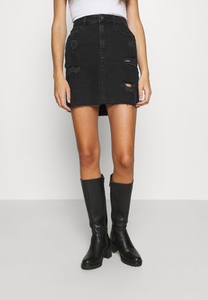 Donna che indossa una minigonna in denim nero consumato, top bianco e stivali in pelle neri al ginocchio, in piedi su uno sfondo semplice.