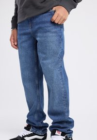 Jeans de mezclilla azul con corte recto, que cuentan con bolsillos y costuras claras. Llevados con zapatillas negras y blancas.