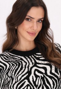 Sieviņa ar gariem brūniem matiem, ģērbusies melnbaltā zebrapatternētā džemperī ar mazām sudraba pērlītēm, maigi smejoties uz gaiša fonda.