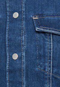 Sinine teksariidest kangas, kus on nähtavad õmblused, hõbedased metallist nupud ja rinnatasku detail. Kulumisega tekstuur ja klassikaline disain.