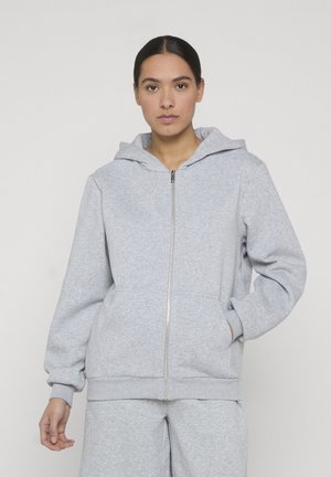 Jonge vrouw met donker haar in een lage knot, gekleed in een lichtgrijze hoodie met rits en bijpassende joggingbroek, tegen een effen achtergrond.