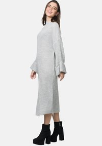 Langes, hellgraues gestricktes Kleid mit Glockenärmeln und seitlichen Schlitzen, kombiniert mit schwarzen Plattform-Stiefeletten. Glattes, strukturiertes Gewebe.