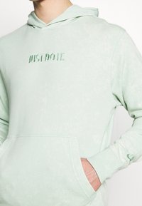 Persona che indossa un hoodie verde chiaro con la scritta "JUST DO IT." ricamata sul petto e con una mano nella tasca frontale, tagliato al collo e in vita.