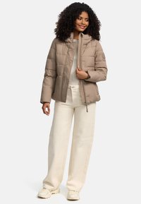 Veste rembourrée beige avec un col montant et une fermeture éclair. Présente une texture lisse et un design matelassé. Associée à un pantalon et des chaussures de couleur claire.