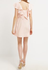 Světle růžové šaty s volánovými rukávy, otevřeným zadním dílem a vypasovaným pasem. Hladká tkanina a délka nad kolena. Nosené s tělovými podpatky.