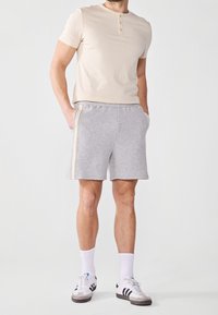 Chemise henley beige clair à manches courtes assortie à un short gris avec des bandes latérales. Portée avec des chaussettes blanches et des baskets grises.
