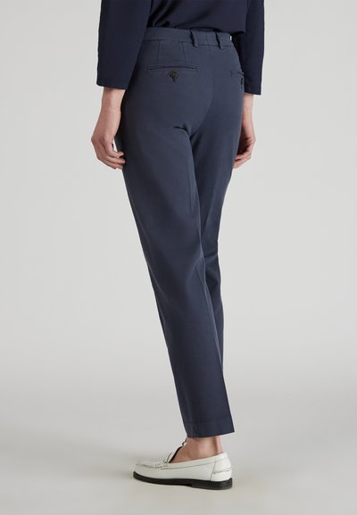 Marineblauwe broek met een getailleerde pasvorm, twee achterzakken, knopenaccenten en een gladde textuur. Gedragen met witte loafers.