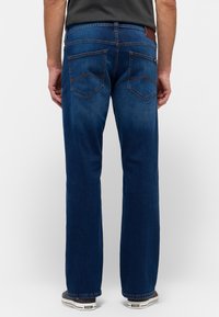 Pantalones vaqueros de mezclilla en azul oscuro, diseño de pierna recta. Presenta dos bolsillos traseros con costuras angulares y un pequeño parche de cuero marrón en la cinturilla.