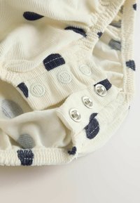 Krēmkrāsas audums ar ribotu struktūru, rotāts ar tumši zilām punktiņām un aizdari ar spiedpogām. Mīkstas, savāktas malas ciešai pieguļai.