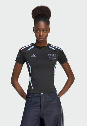 Jonge vrouw met afro-haarstijl, gekleed in een zwart Adidas sportshirt met Audi- en Revolut-logo's, staand met haar handen achter haar rug.