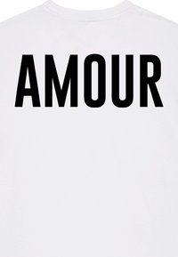 Bijela majica s velikim, podebljanim crnim tekstom na leđima koji glasi "AMOUR". Ima okrugli izrez i kratke rukave.