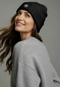 Black ribbed knit beanie with a flat logo patch. Soft texture, fitted design. Worn with a gray sweater, displaying relaxed fit and casual style.