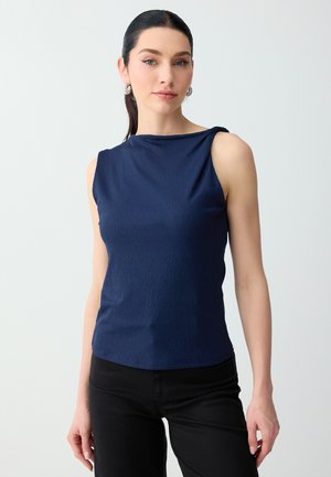 Vrouw met donker haar in een paardenstaart, die zilveren creolen draagt, een mouwloos marineblauw topje en zwarte broek, staand tegen een effen lichte achtergrond.