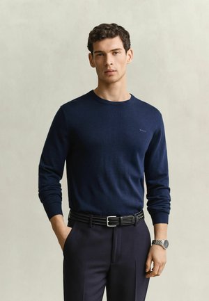 Jeune homme aux cheveux courts et bouclés portant un pull bleu marine, une ceinture noire, un pantalon sombre et une montre-bracelet argentée, debout avec une main dans la poche.