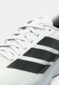 Κοντινή λήψη λευκού αθλητικού παπουτσιού με μαύρες ρίγες και λευκά κορδόνια σε φωτεινό φόντο.