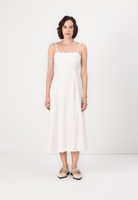 Abito midi bianco con scollo a scalloped, spalline sottili e gonna fluida. Indossato con scarpe beige con dettagli decorativi in metallo.