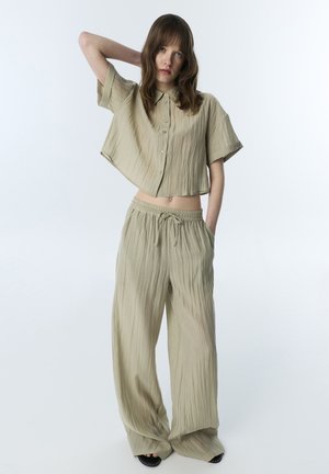 Jonge vrouw in losse, beige gekreukte stoffen outfit met korte, geknoopte blouse en wijde broeken, staand met één hand achter het hoofd.
