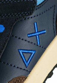 Primo piano di una sneaker in pelle blu navy con forme geometriche ricamate in blu e suola texturizzata beige.