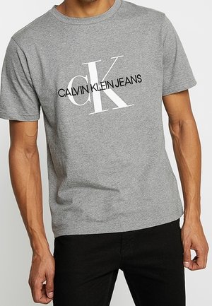 Man draagt een grijs T-shirt met korte mouwen met een grote witte "CK" en zwarte "Calvin Klein Jeans" logo op de borst.