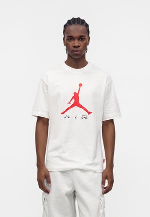 Ung mand med flettet hår iført hvid t-shirt med rød Jumpman-logo og teksten "AIR", kombineret med lysegrå cargoshorts, står neutralt.