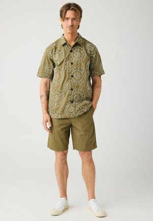 Mand iført en olivengrøn blomstret kortærmet skjorte med knapper, matchende shorts, hvide sneakers, stående med den ene hånd i lommen.