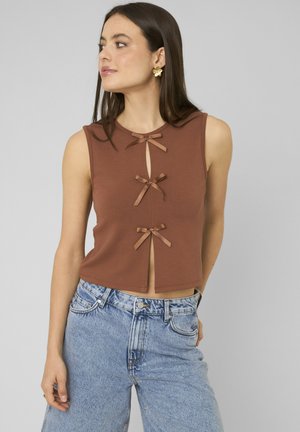 Femme portant un haut marron sans manches avec trois petits nœuds sur le devant et un jean taille haute bleu clair, regardant sur le côté.