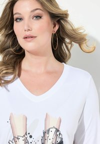 Weißes V-Ausschnitt-Shirt mit einem grafischen Druck von Händen und künstlerischen Details. Das Model hat schulterlanges, welliges Haar und große Creolen.