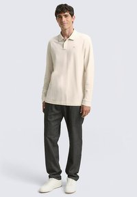 Lichtbeige poloshirt met lange mouwen en een kraag, gedragen met donkergrijze broek en witte sneakers. Heeft een subtiel logo op de linkerborst.