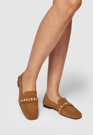 Mocassins en daim marron à semelle plate, avec une bride décorative ornée de clous métalliques sur le devant et des côtés ouverts.