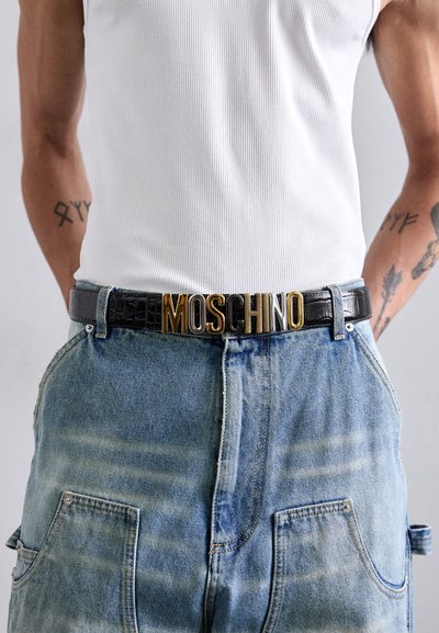 Čierny opasok z kože s embosovaným krokodílím vzorom a zlatou prackou s logom "MOSCHINO", nosený cez modré denimové nohavice s bielym rebrovaným tielkom.