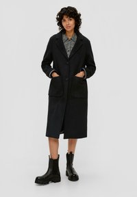 Schwarzer Wollmischungs-Mantel mit Knopfverschluss, großen Fronttaschen und kontrastierenden inneren Bündchen. Kombiniert mit klobigen schwarzen Stiefeln bis zum Knöchel.