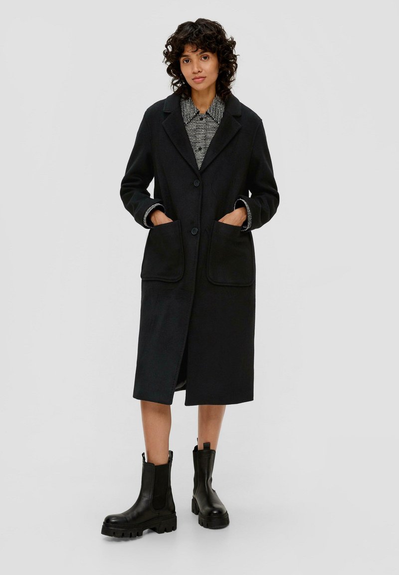 Schwarzer Wollmischungs-Mantel mit Knopfverschluss, großen Fronttaschen und kontrastierenden inneren Bündchen. Kombiniert mit klobigen schwarzen Stiefeln bis zum Knöchel.