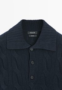 Pull en laine bleu marine avec un col côtelé, cinq boutons noirs et un motif en torsade. L'étiquette comprend la marque et les informations d'entretien.