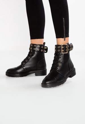 Czarne skórzane botki na kostkę ze złotymi oczkami, sznurowane z przodu i szerokimi paskami z klamrami, noszone z czarnymi legginsami z bocznymi zamkami.