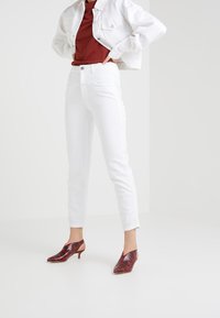 Mujer usando jeans blancos cortos, zapatos de tacón bajo con patrón de serpiente roja, camisa rojo óxido y chaqueta blanca corta, de pie con las manos en las caderas.