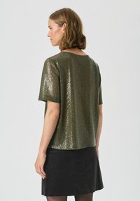 Top scurt cu mânecă scurtă de culoare verde măsliniu, decorat cu paiete, cu o croială lejeră și guler rotund, asortat cu o fustă neagră, având o suprafață texturată.